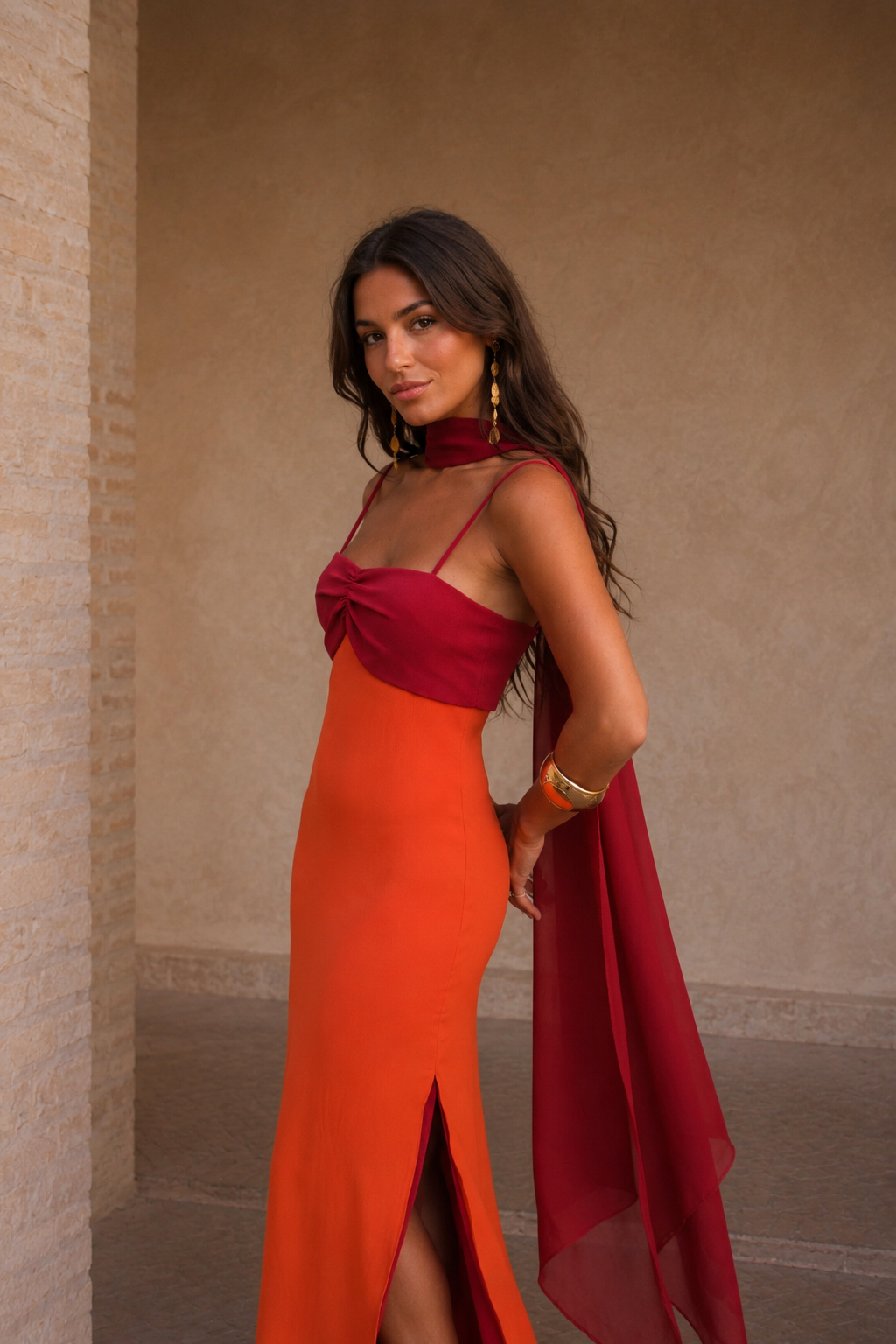 Robe longue orange fendue, buste bordeaux noué, voile fluide drapé sur l’épaule.