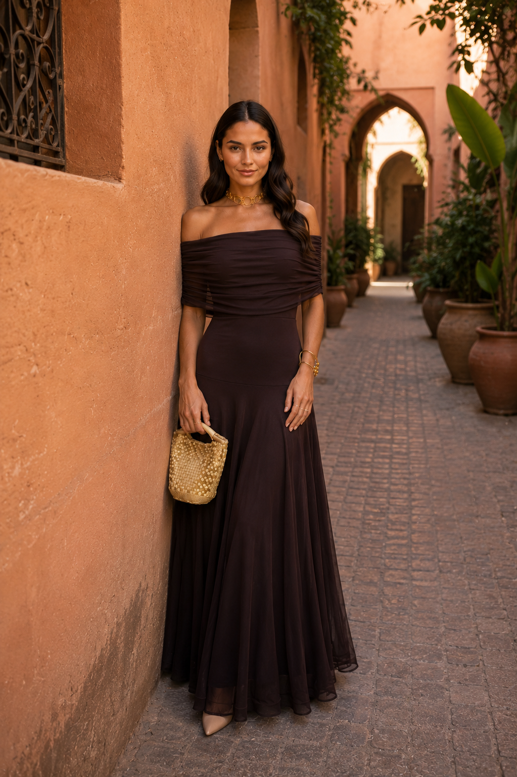 Robe longue marron foncé à drapé, épaules dénudées, pochette dorée à la main.