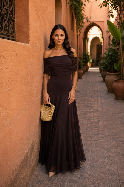 Robe longue marron foncé à drapé, épaules dénudées, pochette dorée à la main.