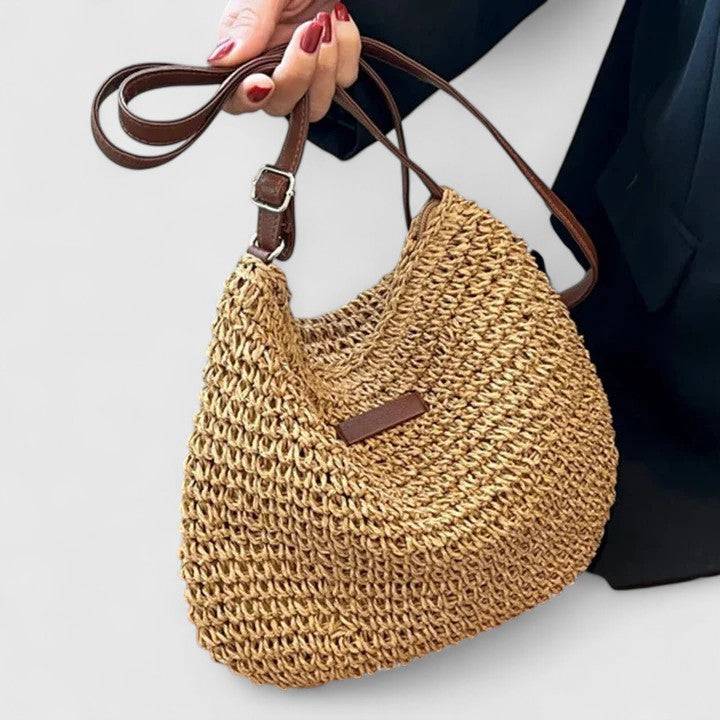 Sac en paille beige porté à la main, anse marron, texture tressée et patch rectangulaire.